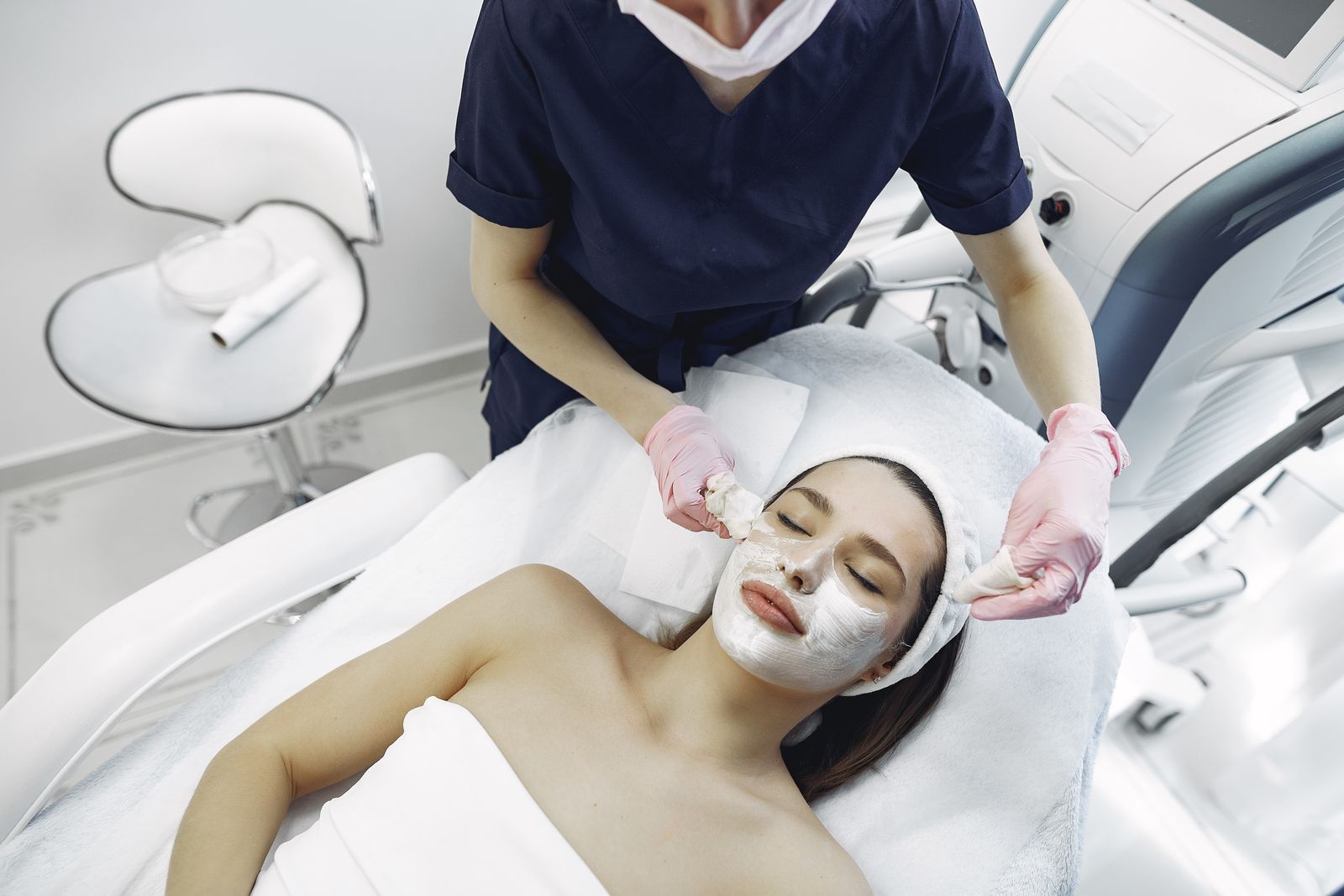 Esthetician in gloves removing a purifying facial mask from a relaxed client